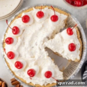 Overhead view of a Million Dollar Pie in a graham cracker crust topped with dollops of whipped cream and maraschino cherries, with one slice removed.