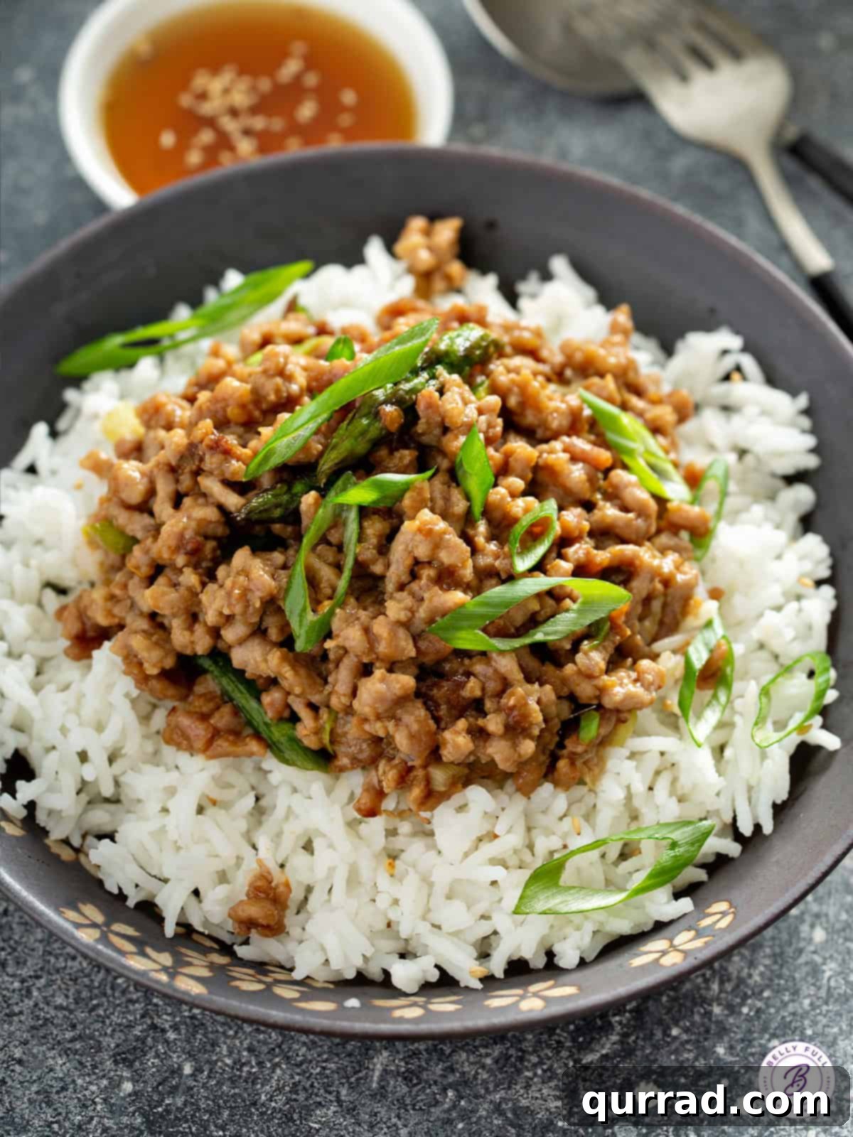 A vibrant and saucy ground pork stir fry, garnished with fresh green scallions, artfully presented over white rice in a bowl, ready for enjoyment.