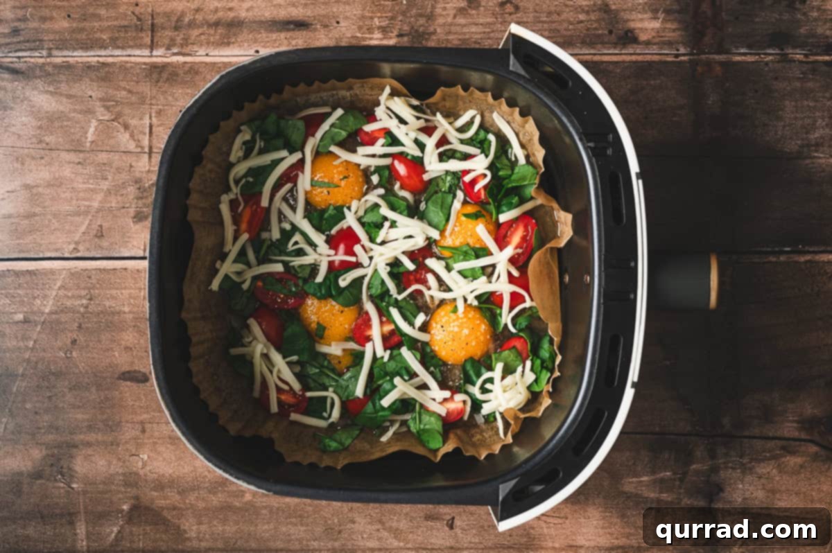 Fluffy Air Fryer Egg Frittata 5 Eggs in the air fryer basket topped with vibrant halved cherry tomatoes, fresh chopped spinach, and a generous layer of shredded mozzarella cheese, ready for cooking.