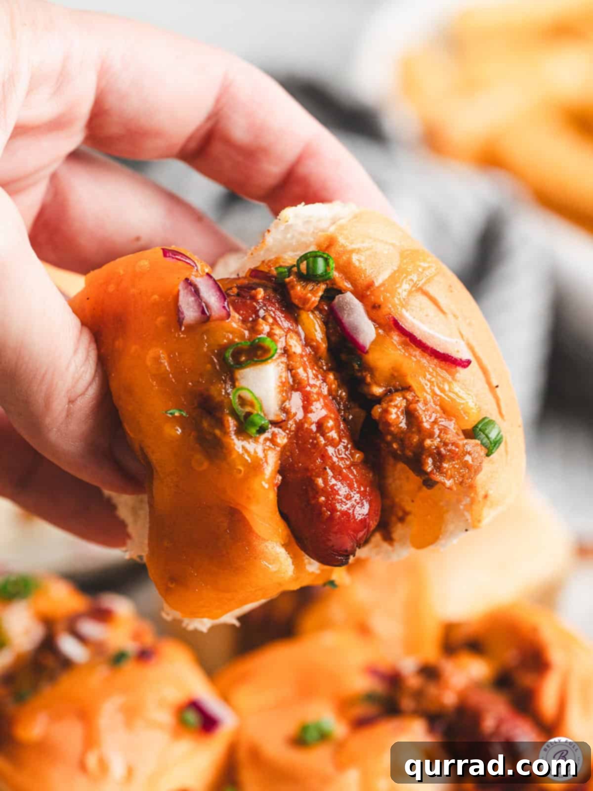Bite-Sized Chili Cheese Dogs 2 Close-up of a hand holding a mini chili cheese dog with melted cheddar, chili, diced red onion, and scallions on a Hawaiian roll, showcasing its delicious layers and perfect bite-size.