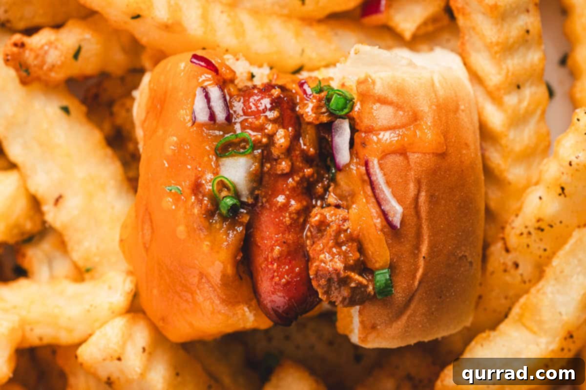 Bite-Sized Chili Cheese Dogs 8 A mini chili cheese dog on a plate, surrounded by seasoned crinkle-cut fries, topped with hearty chili, melted cheddar, red onions, and green scallions, showcasing a complete snack.