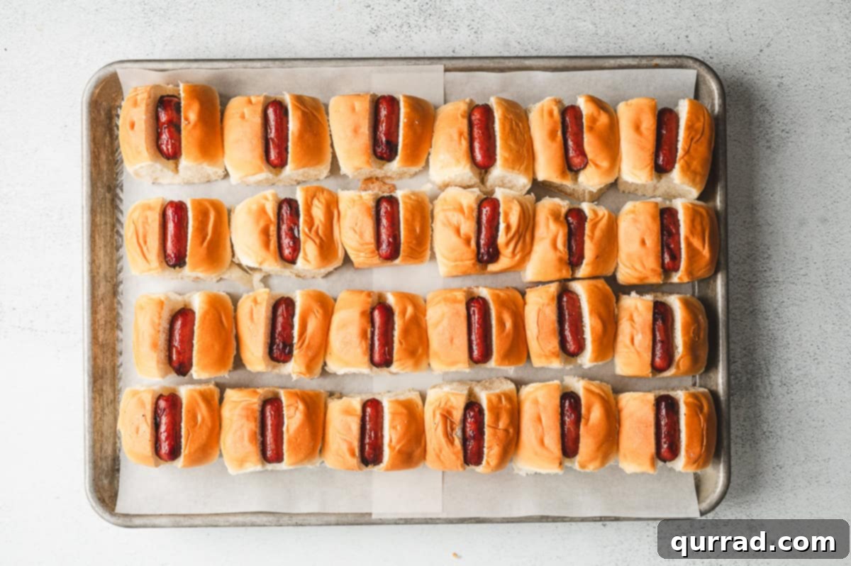 Bite-Sized Chili Cheese Dogs 4 Cocktail franks neatly tucked into the sliced Hawaiian rolls, arranged on a parchment-lined baking sheet, ready for the next delicious layer.