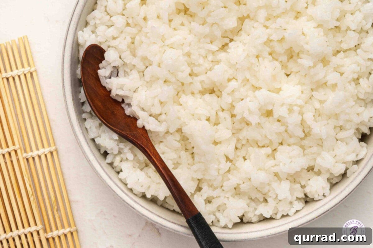Secrets to Perfect Sushi Rice 9 Close-up of glossy sushi rice in a ceramic bowl with a wooden spoon, next to a bamboo sushi mat.