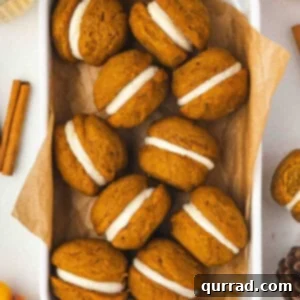 A white dish lined with parchment paper filled with pumpkin whoopie pies sandwiched with cream cheese frosting, surrounded by cinnamon sticks and fall decorations.