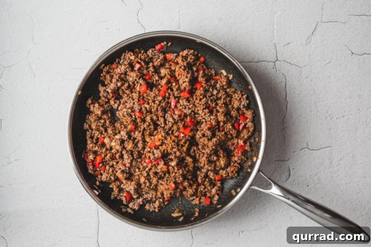 Large skillet filled with browned ground beef, diced onions, and red bell peppers cooked together for the casserole filling.