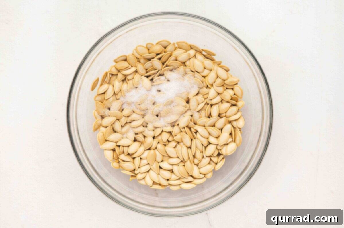 Raw pumpkin seeds in a glass bowl, glistening with olive oil and sprinkled with coarse kosher salt, awaiting a thorough toss.
