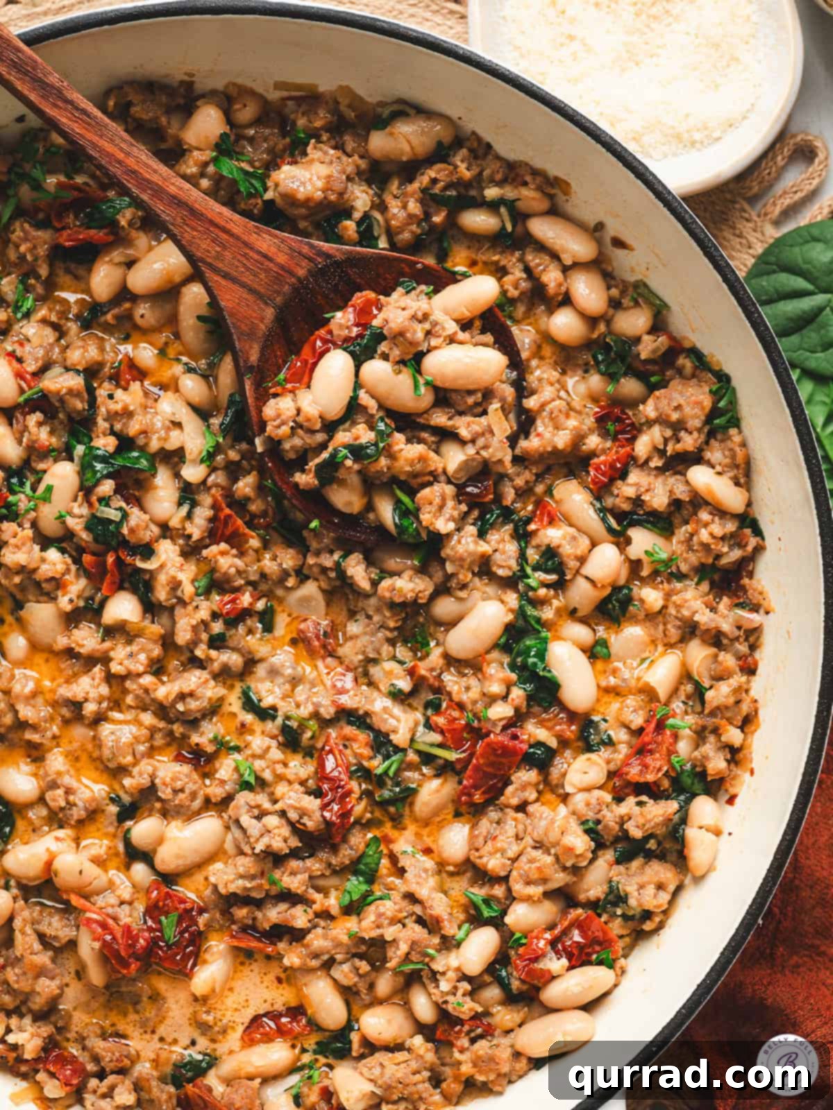 Tuscan Sausage and White Bean Skillet 2 Overhead view of Tuscan white bean and sausage skillet in a pan, with a wooden spoon lifting a serving of beans, sausage, spinach, and sun-dried tomatoes.