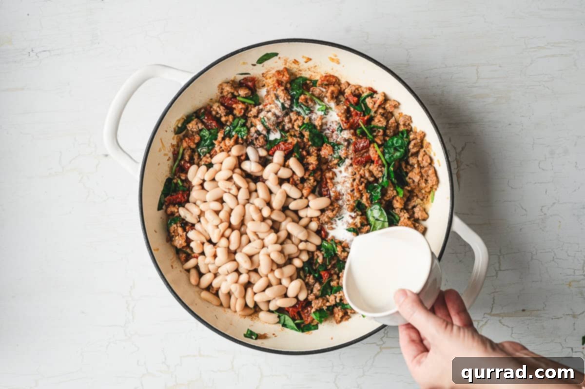 Tuscan Sausage and White Bean Skillet 6 Hand pouring cream into a skillet filled with cooked sausage, spinach, sun-dried tomatoes, and cannellini beans.
