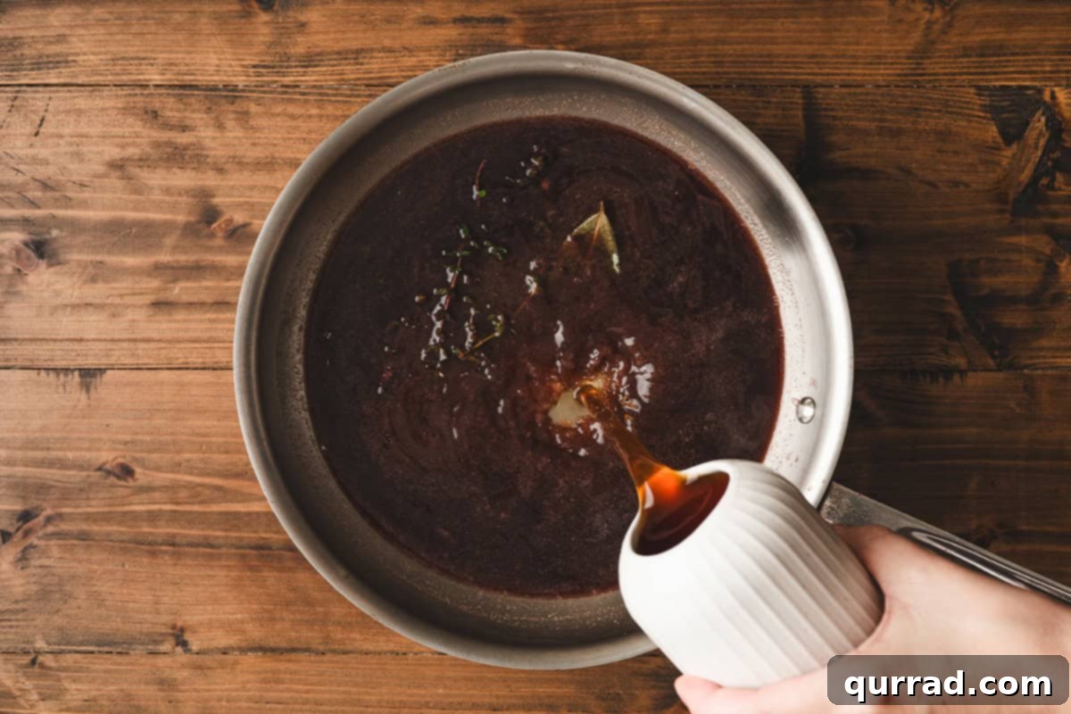 Velvety Red Wine Glaze 7 Beef stock being poured into the simmering red wine and herb mixture in a stainless steel skillet.