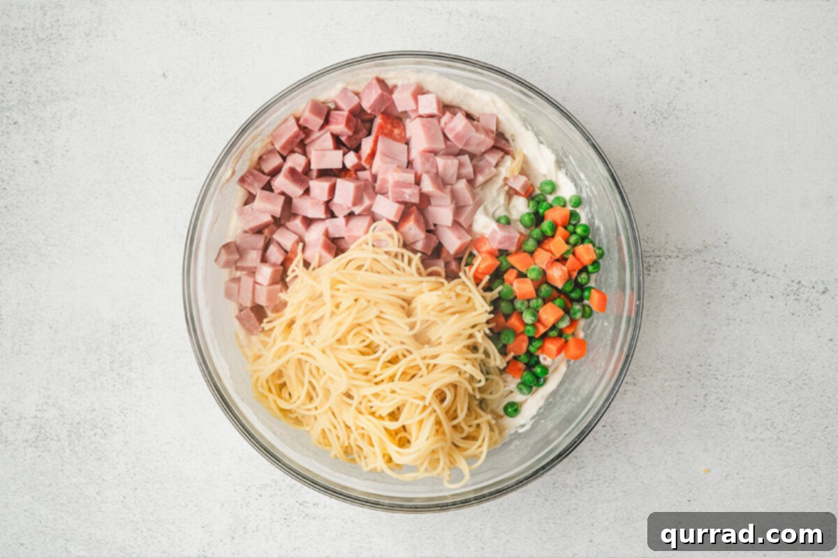 Rustic Tetrazzini 7 Cooked spaghetti, diced ham, and frozen peas and carrots being stirred into the creamy sauce in a large bowl.