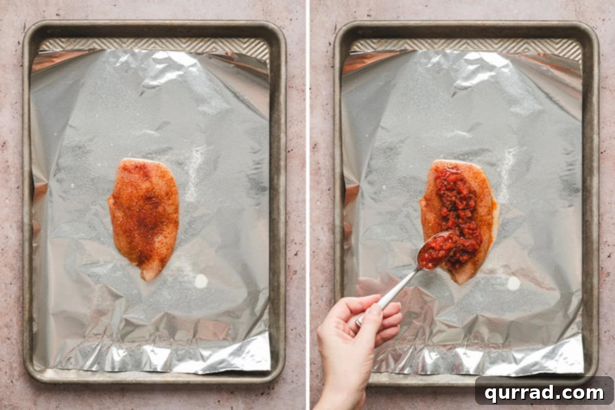 A seasoned chicken breast being topped with zesty salsa on a foil sheet.