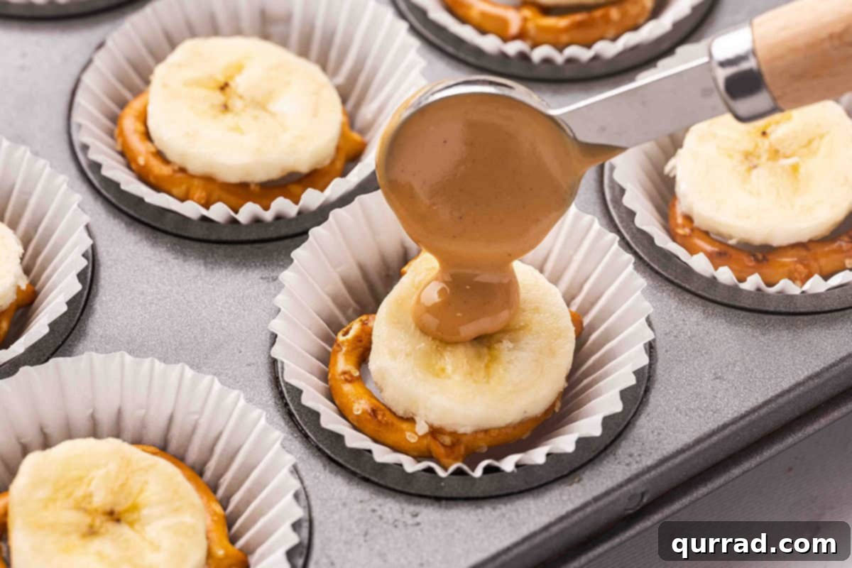 A spoon drizzling warmed peanut butter over a banana slice and pretzel in a muffin liner.
