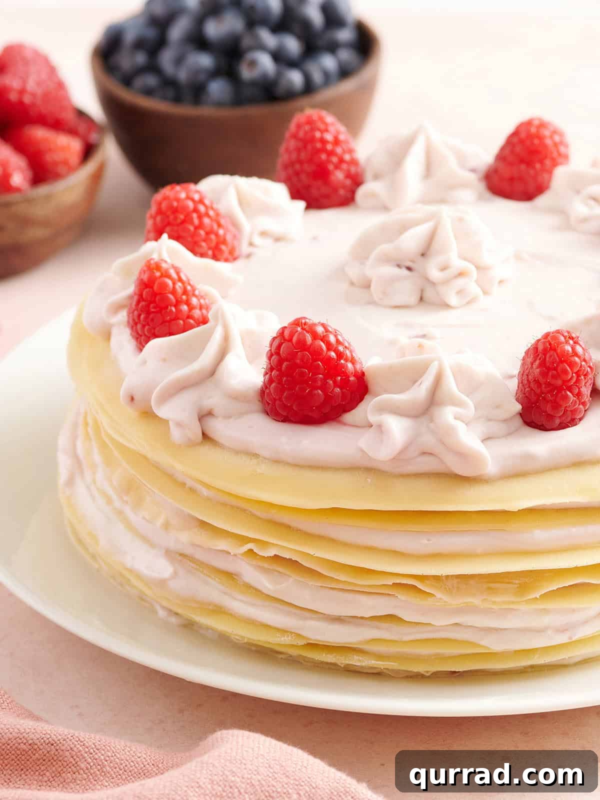 How to Make a Classic French Crepe Cake (Mille Crêpes) 2 A layered crepe cake filled with raspberry cream, topped with piped cream swirls and fresh raspberries, with bowls of blueberries and raspberries in the background.