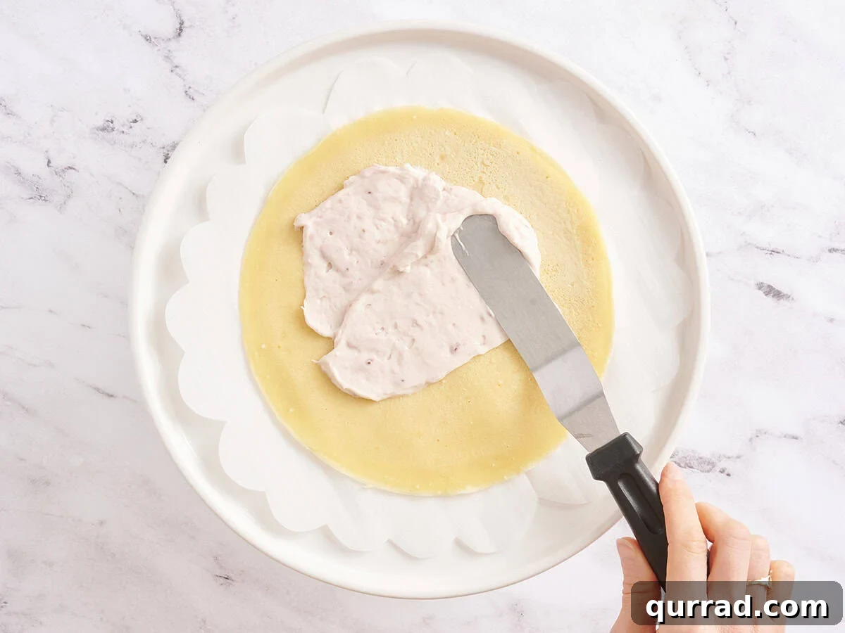 How to Make a Classic French Crepe Cake (Mille Crêpes) 10 Spreading raspberry cream filling over a single crepe layer with an offset spatula on a parchment-lined plate.