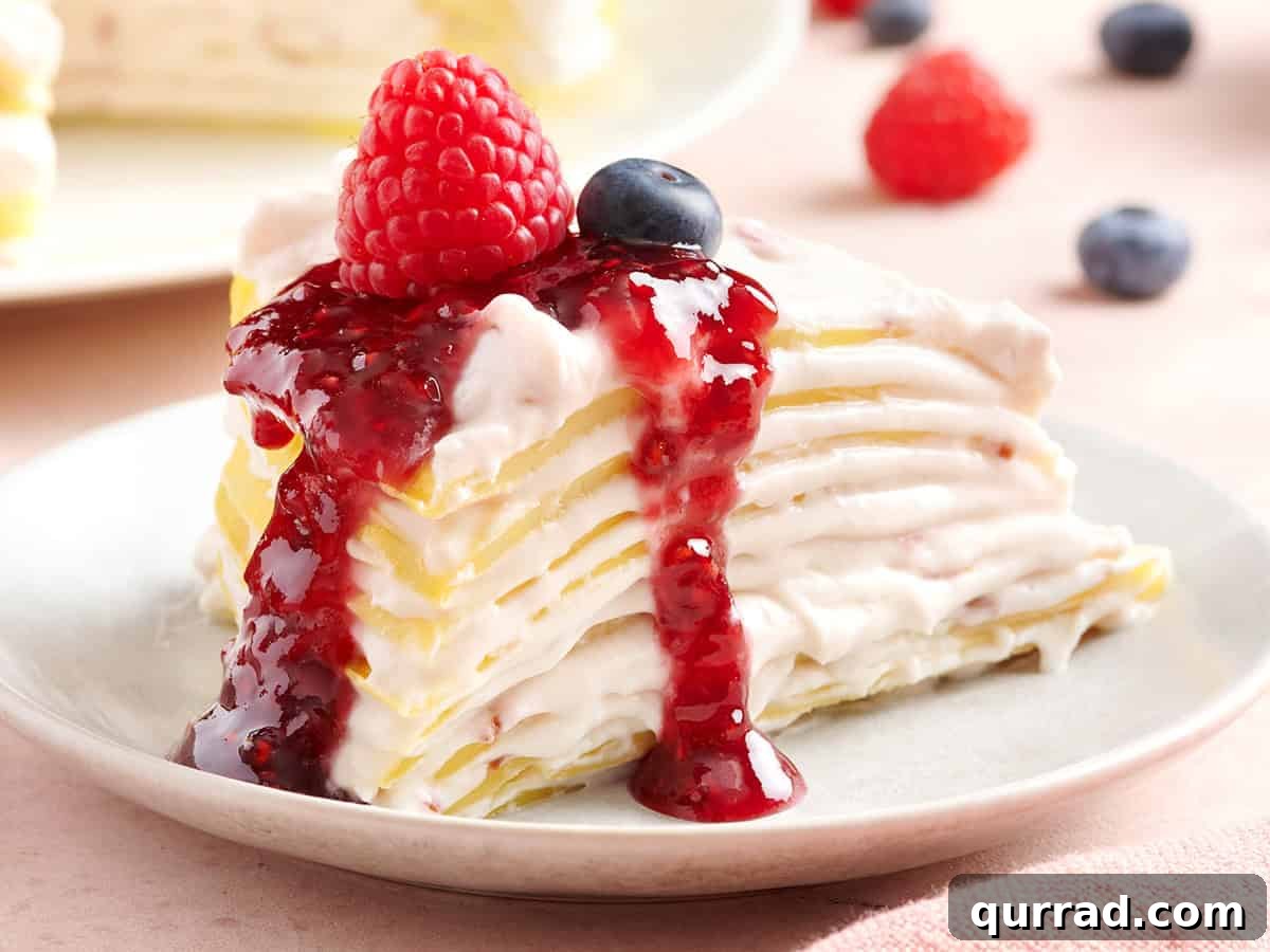 How to Make a Classic French Crepe Cake (Mille Crêpes) 11 A slice of crepe cake topped with raspberry jam sauce, fresh raspberry, and blueberry on a white plate, with more berries scattered in the background.