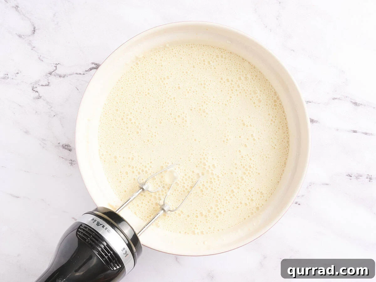 How to Make a Classic French Crepe Cake (Mille Crêpes) 6 A mixing bowl of smooth crepe batter after whisking with a hand mixer, resting on a white marble surface.