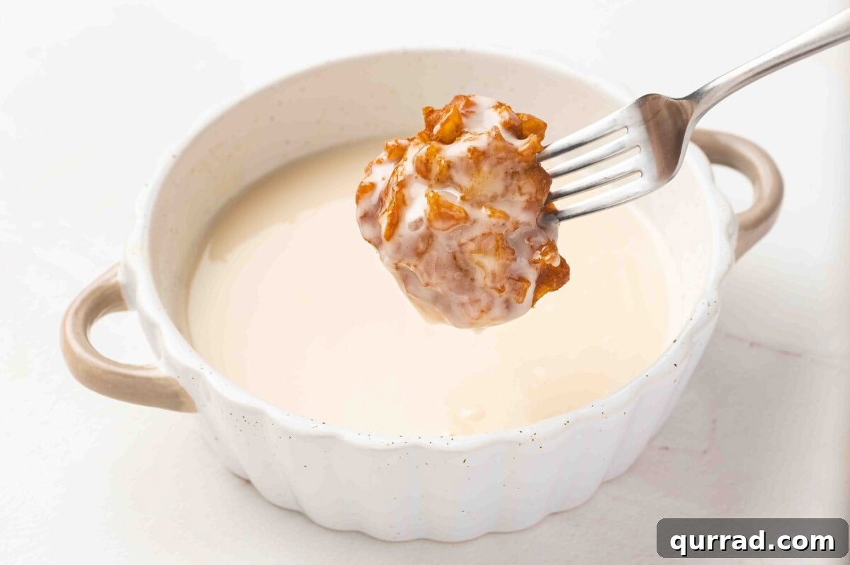 A golden apple fritter held with a fork being dipped into a bowl of vanilla glaze.