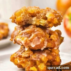 A stack of three glazed apple fritters on a white plate, the top one with a bite taken out, showing chunks of apple inside.